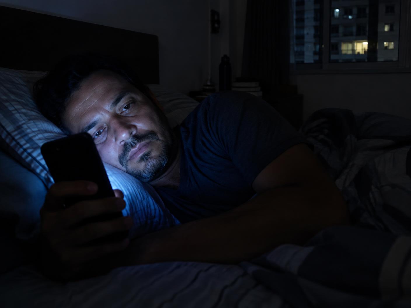 Homem na cama à noite com o rosto iluminado pela tela do celular